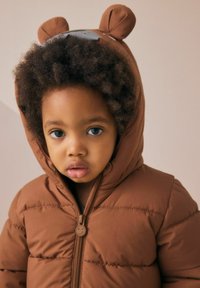 Brązowa pikowana kurtka z kapturem w kształcie misia, z pluszowymi uszami i suwakiem z uśmiechniętą twarzą, o teksturowanej powierzchni i pikowanym wzorze.