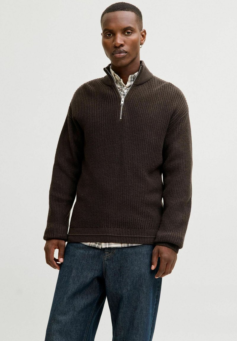 Dunkelbrauner gestrickter Pullover mit einem Halbreißverschluss-Kragen, strukturiertem Gewebe und lockerer Passform, über einem karonierten Hemd getragen. Denim-Jeans sind am unteren Rand sichtbar.