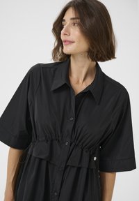 Abito chemisier nero con maniche corte, caratterizzato da una vita arricciata, orlo volants e una texture di tessuto liscia.