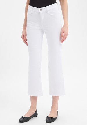 Vrouw die witte cropped flare jeans en zwarte balletschoenen draagt, staand tegen een effen witte achtergrond.