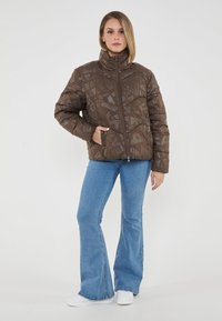 Giacca a vento marrone con motivo geometrico, collo alto e chiusura a zip, abbinata a jeans blu a zampa d'elefante e sneakers bianche.