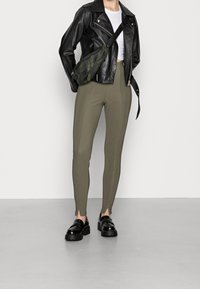 Osoba w czarnej skórzanej kurtce, białym topie na krótki rękaw, oliwkowych dopasowanych spodniach z przodu z rozcięciami, czarnych masywnych loafersach i torbie na ramię w kamuflażu.