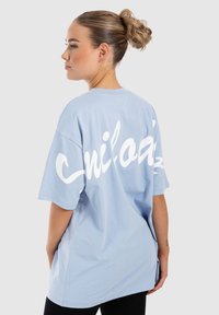 Camiseta de gran tamaño de color azul claro con mangas cortas, que presenta un gran texto gráfico en cursiva blanca en la parte posterior. Textura de algodón suave.