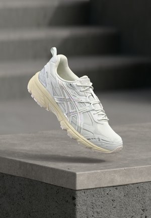 Chaussure de sport gris clair avec tige en mesh, accents réfléchissants et semelle en caoutchouc. Présente un design à lacets et un col rembourré.