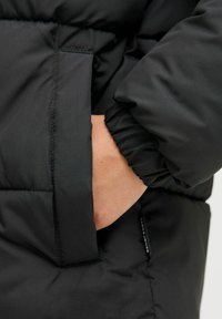 Detaljer av en svart puffjakke med løs passform, elastiske mansjetter og en sidelomme. Materialet virker glatt og litt blankt.