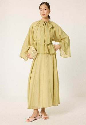 Femme portant une longue robe jaune clair à larges manches et col noué, tenant une pochette tissée et chaussant des chaussures ouvertes rose.
