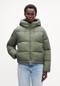 Olivenfarget pufferjakke med hette, strukturert stoff, glidelås, og en liten logo; har sydd seksjoner og en noe oversized design.