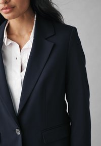 Ruisikankainen tummansininen blazer, jossa on jämäkkä muotoilu, yksittäinen napitus, läppäkaulus ja ommellut olkahaituvat, valkoisen paidan päällä.