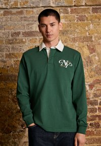 Polo shirt vert à manches longues avec un col blanc, doté d'un logo brodé et de détails complexes. Porté avec un jean en denim foncé.