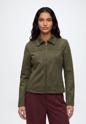 Frau mit langen dunklen Haaren trägt eine olivgrüne Jacke mit Reißverschluss und bordeauxrote Hosen und steht vor einem schlichten hellen Hintergrund.