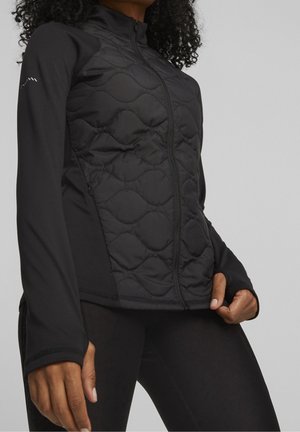 Mujer con una chaqueta negra acolchada con cremallera, mangas largas y orificios para los pulgares, posando con una mano tirando del dobladillo de la chaqueta.