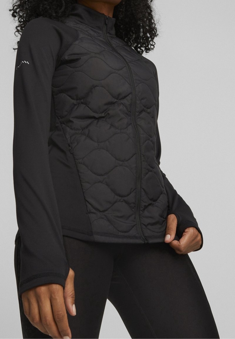 Femme portant une veste matelassée noire zippée, manches longues avec trous pour les pouces, posant avec une main tirant l'ourlet de la veste.