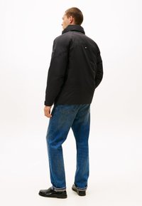 Sort pufferjakke med høj krave, med lynlås og logoaccent. Sammen med blå jeans og sorte sko.