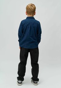 Cămașă albastră cu butoni, cu mâneci lungi, pliuri pe spate și blugi negri. Adidași negri cu accente albe. Pe un fundal neutru.