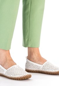 Weiße durchbrochene Slipper mit einem gemusterten Design und brauner Gummisohle, kombiniert mit hellgrünen, knöchellangen Hosen.