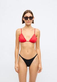Κόκκινο μπικίνι με κυμματιστές άκρες, ρυθμιζόμενες τιράντες και μαύρο σλιπάκι, με απλή υφή και μινιμαλιστικό σχέδιο. Μαύρα γυαλιά ηλίου.