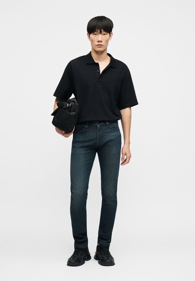 Μαύρο πόλο μπλουζάκι με δύο κουμπιά, στενά σκουρόχρωμα μπλε τζιν και μαύρα αθλητικά παπούτσια. Το μοντέλο κρατά μια μαύρη τσάντα, στέκεται μπροστά σε απλό φόντο.