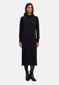 Robe noire à manches longues avec un col haut, fabriquée dans un tissu doux et texturé. La robe est longue jusqu'aux chevilles avec une silhouette droite.