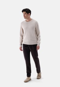 Maglione beige con polsini e orlo a costine, indossato con pantaloni in velluto a coste scuri e stivaletti beige. Il modello è in piedi su uno sfondo bianco.