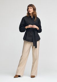 Czarna koszula zapinana na guziki z przewiązaniem w talii, z bufiastymi rękawami; połączona z beżowymi spodniami o szerokich nogawkach i brązowymi butami na obcasie.