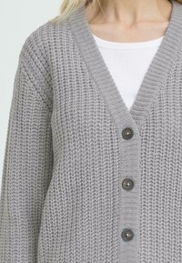 Cárdigan gris de punto con escote en V, mangas largas y tres botones en la parte delantera. Tejido texturizado con un corte holgado, diseño casual.