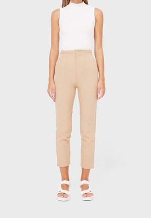 Femme portant un pantalon beige taille haute court, un top blanc sans manches à col montant factice, et des sandales blanches ouvertes à l'avant avec des brides à la cheville, debout.