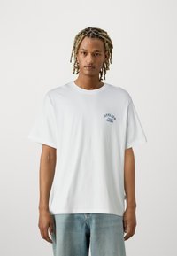 Άσπρο βαμβακερό μπλουζάκι με χαλαρή εφαρμογή, διαθέτει μπλε γραφικό κείμενο "ATELIER" στο αριστερό στήθος. Κοντά μανίκια, στρογγυλή λαιμόκοψη.