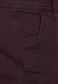 Burgundy-huokoset kangashousut, joissa on sileä pinta, kulmalistalla varustetut etutaskut ja erottuvat ompeleet saumoissa.