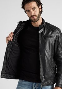 Schwarze Lederjacke mit gesteppten Schulterdetails, Reißverschluss vorne und einer kleinen Brusttasche. Das Model trägt ein schwarzes Shirt darunter.