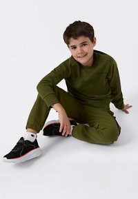 Pojke med kort brunt hår som ler, sitter på golvet klädd i en olivgrön sweatshirt och byxor, vita strumpor och svarta sneakers med orange detaljer.