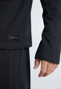 Chemise noire à manches longues en tissu doux, avec un ourlet arrondi, des coutures visibles et une étiquette logo Diesel sur le côté.