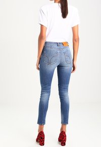 Vaqueros de corte ajustado en denim azul con cintura de altura media, cinco bolsillos, un parche de cuero en la parte posterior y textura desgastada. Combinados con tacones de leopardo rojo.