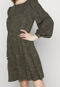 Robe vert olive avec imprimé léopard noir, manches longues bouffantes, encolure ronde et bas évasé, avec un taille ajustée et des poignets boutonnés.