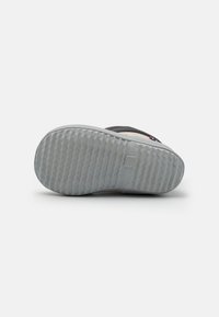 Semelle d'une chaussure de sport avec une surface en caoutchouc gris texturé, dotée d'un motif ondulé et d'un petit logo en relief au niveau du talon.