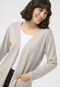 Beige langekleed cardigan met V-hals, vervaardigd uit zachte stof. Heeft een open voorkant en twee voorzakken. Lichtgewicht en ontspannen pasvorm.