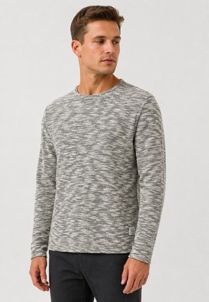 Man die een grijs-wit gemêleerd shirt met lange mouwen en een zwarte broek draagt, kijkt naar links tegen een effen achtergrond.