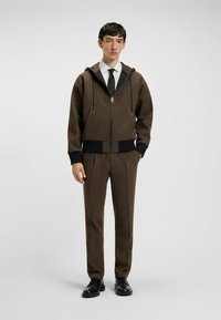 Bruine gestructureerde zip-hoodie met bijpassende broeken, voorzien van een zwarte tailleband, hoekzakken en zwarte schoenen. Een wit overhemd en stropdas maken de look compleet.