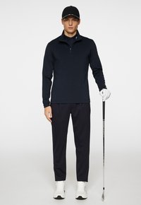 Polo a maniche lunghe blu navy con colletto a mezza zip, abbinato a pantaloni coordinati. Il modello tiene in mano un bastone da golf, indossando scarpe da ginnastica bianche e un cappellino.