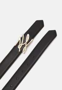 Ceinture en cuir noir avec boucle signature en métal doré. Présente des pointes effilées et plusieurs trous pour l'ajustement. Texture lisse.