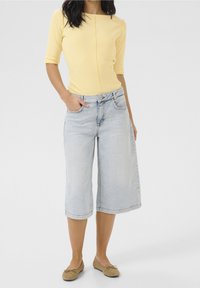 Top giallo aderente con maniche a tre quarti, abbinato a culotte in denim azzurro chiaro e scarpe basse beige, dotato di tasche frontali e una vestibilità rilassata.