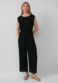 Schwarzer plissierter Jumpsuit mit kurzen Ärmeln, taillierter Passform, weitem Bein und strukturiertem Stoff. Getragen mit flachen Sandalen mit Knoten-Detail.