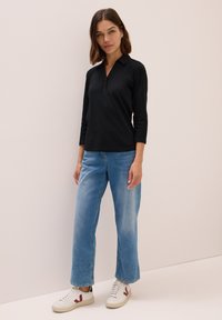 Polo shirt noir avec trois boutons, manches trois-quarts. Jean droit bleu clair et baskets blanches avec des accents rouges. Design simple et épuré.