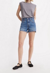 Person med ärmlös grå topp, högmidjade slitna denimshorts och svarta Mary Jane-skor, som står mot en enfärgad vit bakgrund.