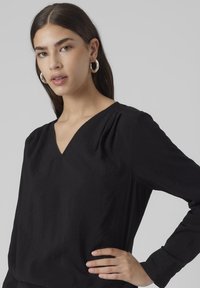 Blusa nera a maniche lunghe con scollatura a V e sottili pieghe sulle spalle, realizzata in tessuto leggero con una texture liscia.