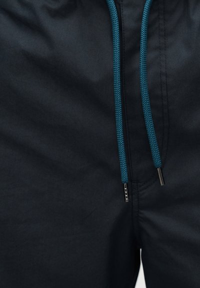 Σκούρα σορτς με τιρκουάζ κορδόνια με μεταλλικά άκρα και ραφές γύρω από τη μέση και την περιοχή του φερμουάρ.