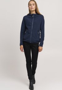 Giacca zip-up in pile blu navy con cappuccio, due tasche con zip, maniche lunghe e design aderente; abbinata a pantaloni neri slim-fit e stivaletti.