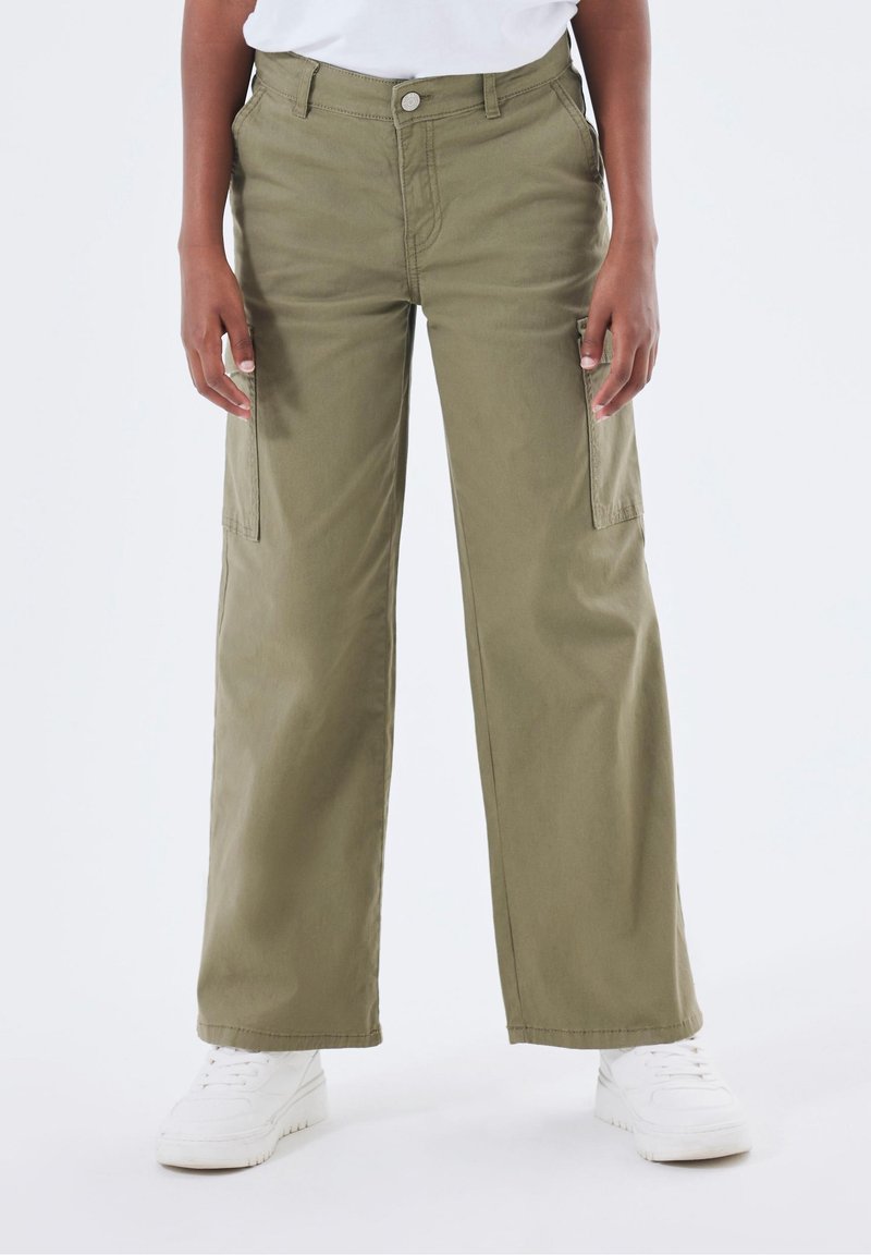 Personne portant un pantalon cargo large olive et des baskets blanches, debout devant un fond clair uni.