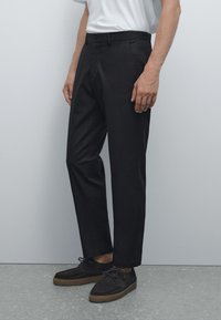 Černé bavlněné kalhoty s rovnou rovnicí, vybavené zapínáním na knoflík a bočními kapsami, doplněné o tmavé semišové boty.