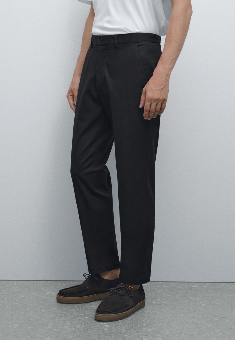 Černé bavlněné kalhoty s rovnou rovnicí, vybavené zapínáním na knoflík a bočními kapsami, doplněné o tmavé semišové boty.