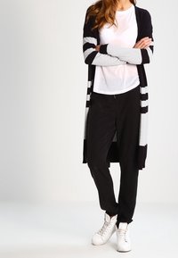 Lange cardigan met zwarte en grijze strepen, wit T-shirt, zwarte broek en witte sneakers. Zachte stof en ontspannen pasvorm ontwerp.